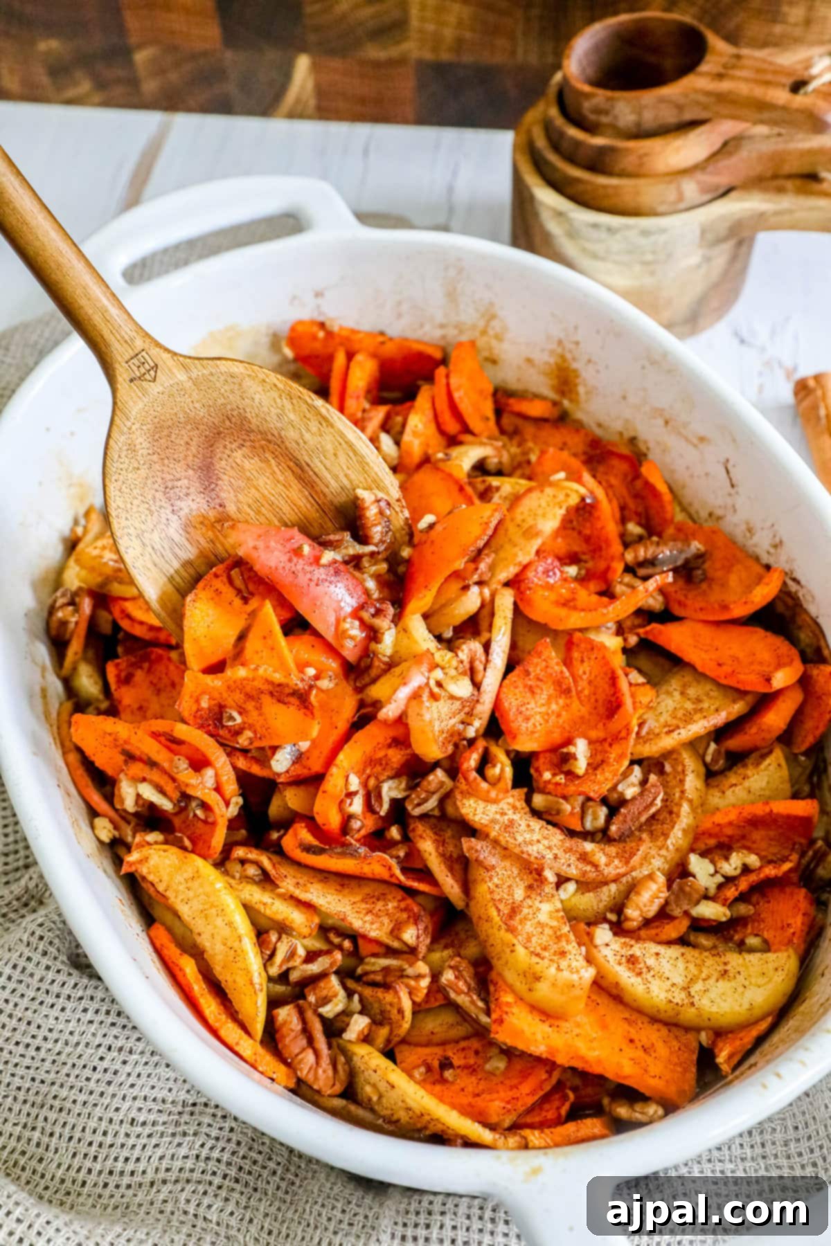 Roasted carrots and pecans in a white dish with a wooden spoon, accompanied by cinnamon sweet potato and apple bake in another dish, all on a wooden surface.