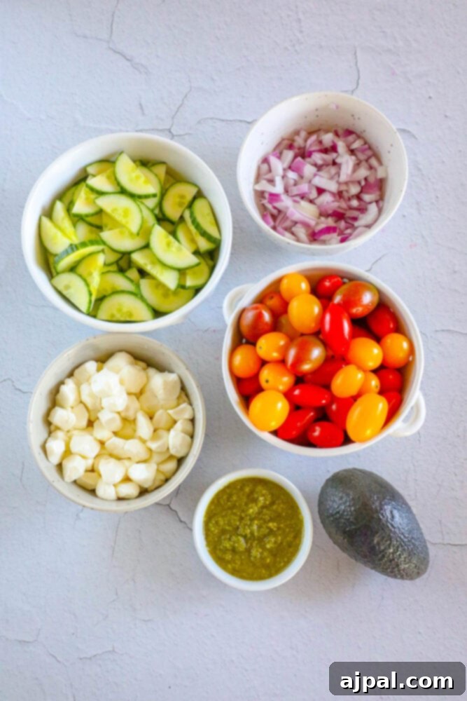 Vibrant Pesto Caprese 5 Sliced cucumbers in a bowl next to bowls of diced onions, cherry tomatoes, crumbled feta, and pesto, with a whole avocado on the table, showcasing all fresh ingredients.