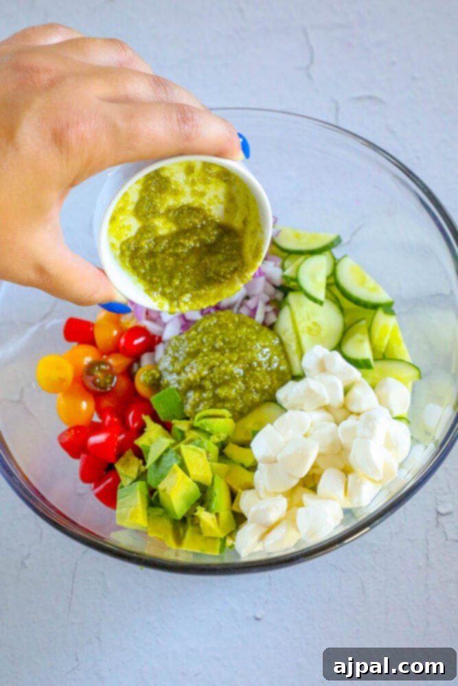 Vibrant Pesto Caprese 7 A hand expertly pouring rich, green pesto sauce over a vibrant medley of chopped vegetables in a bowl, illustrating the dressing process.