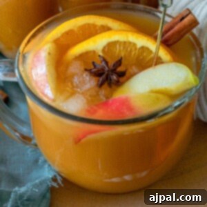 A festive clear mug of pumpkin punch with ice, sliced apples and oranges, a cinnamon stick, and a star anise, perfect for fall celebrations.