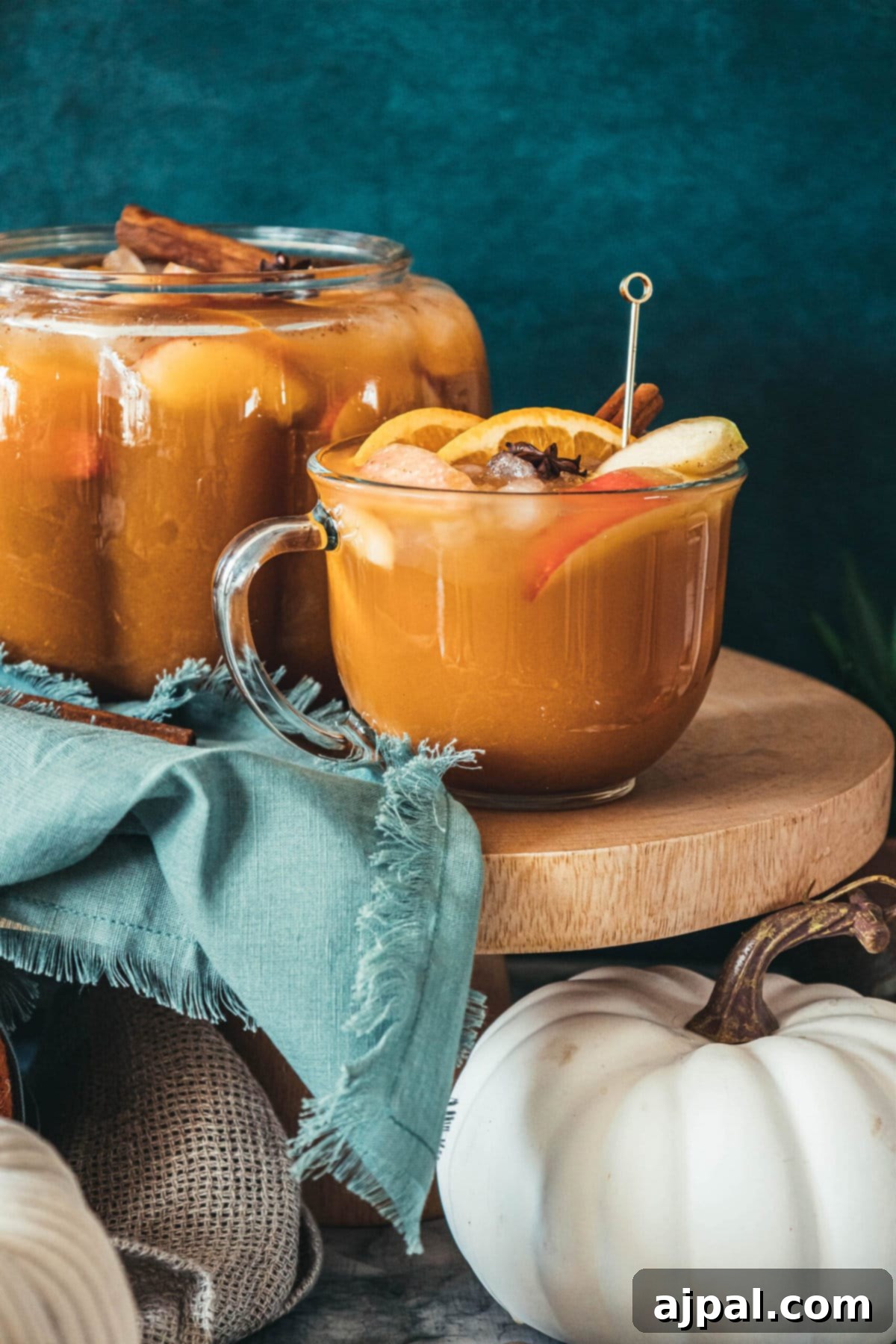 A clear glass of spiced pumpkin cider punch, adorned with cinnamon sticks and apple slices, resting on a rustic wooden table amidst decorative pumpkins, creating a warm fall scene.