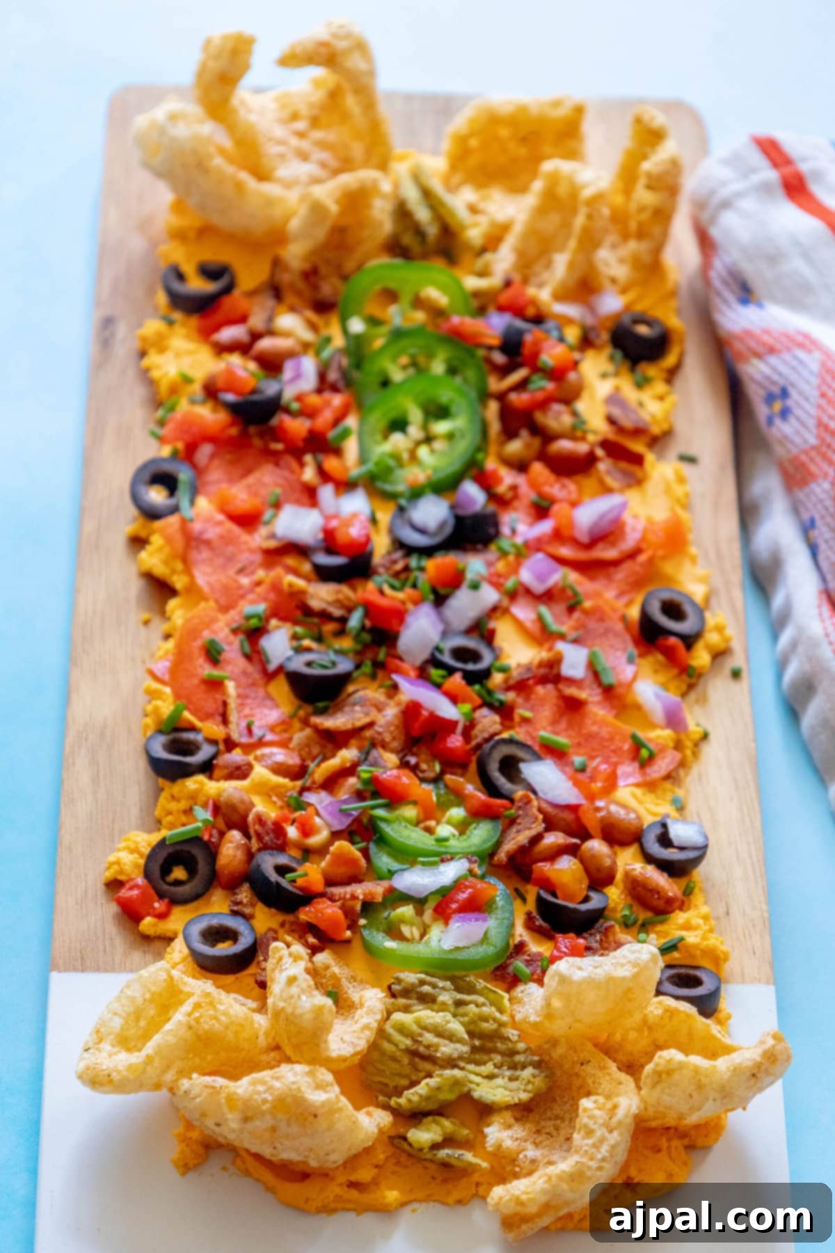 picture of pub cheese spread on a board covered with pork rinds, jalapenos, olives, bacon crumbles, peanuts, pimiento peppers, chives, red onion, and pepperoni