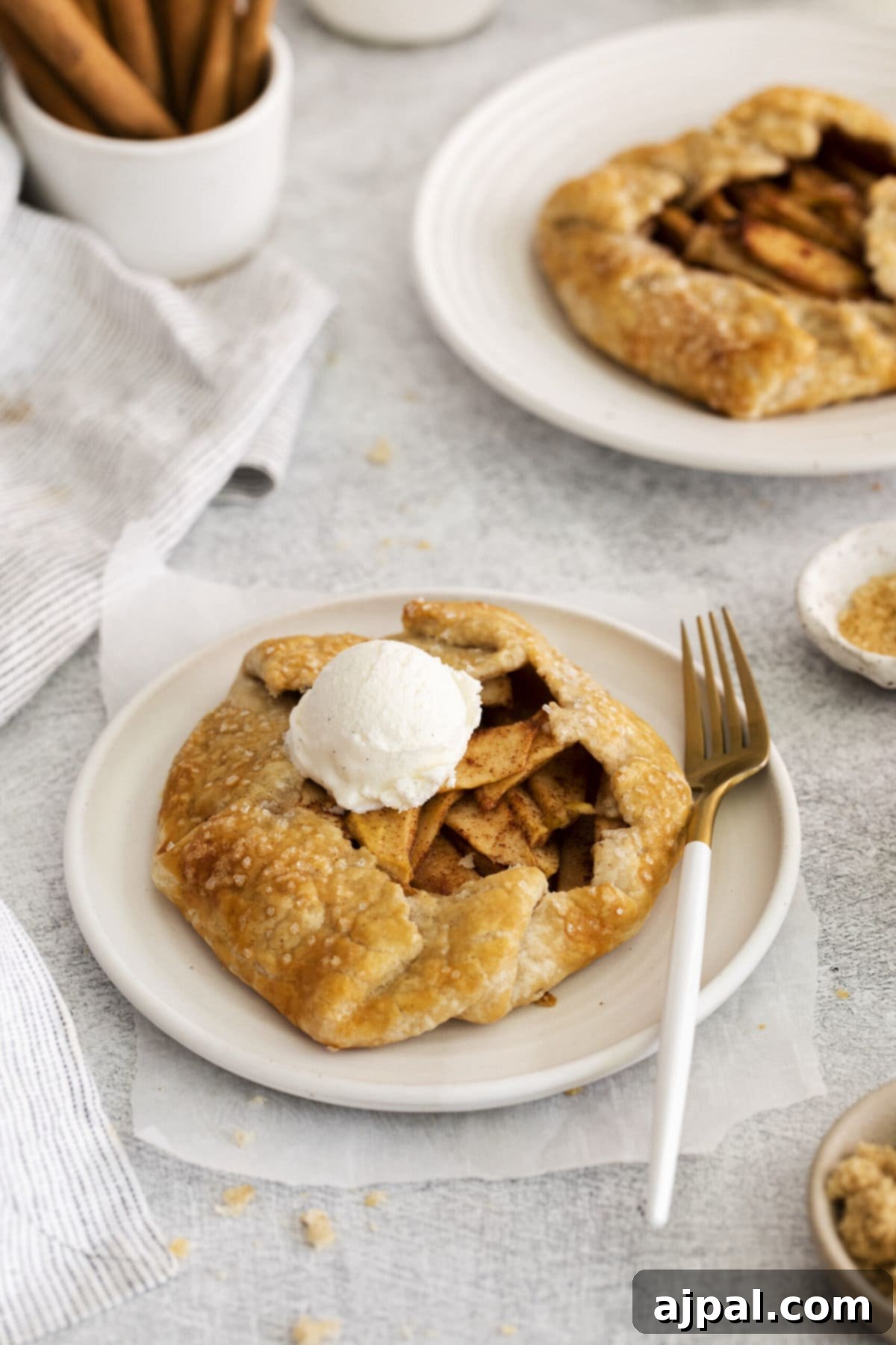Two baked apple galettes, beautifully golden, served with whipped cream and cinnamon sticks on a rustic table.