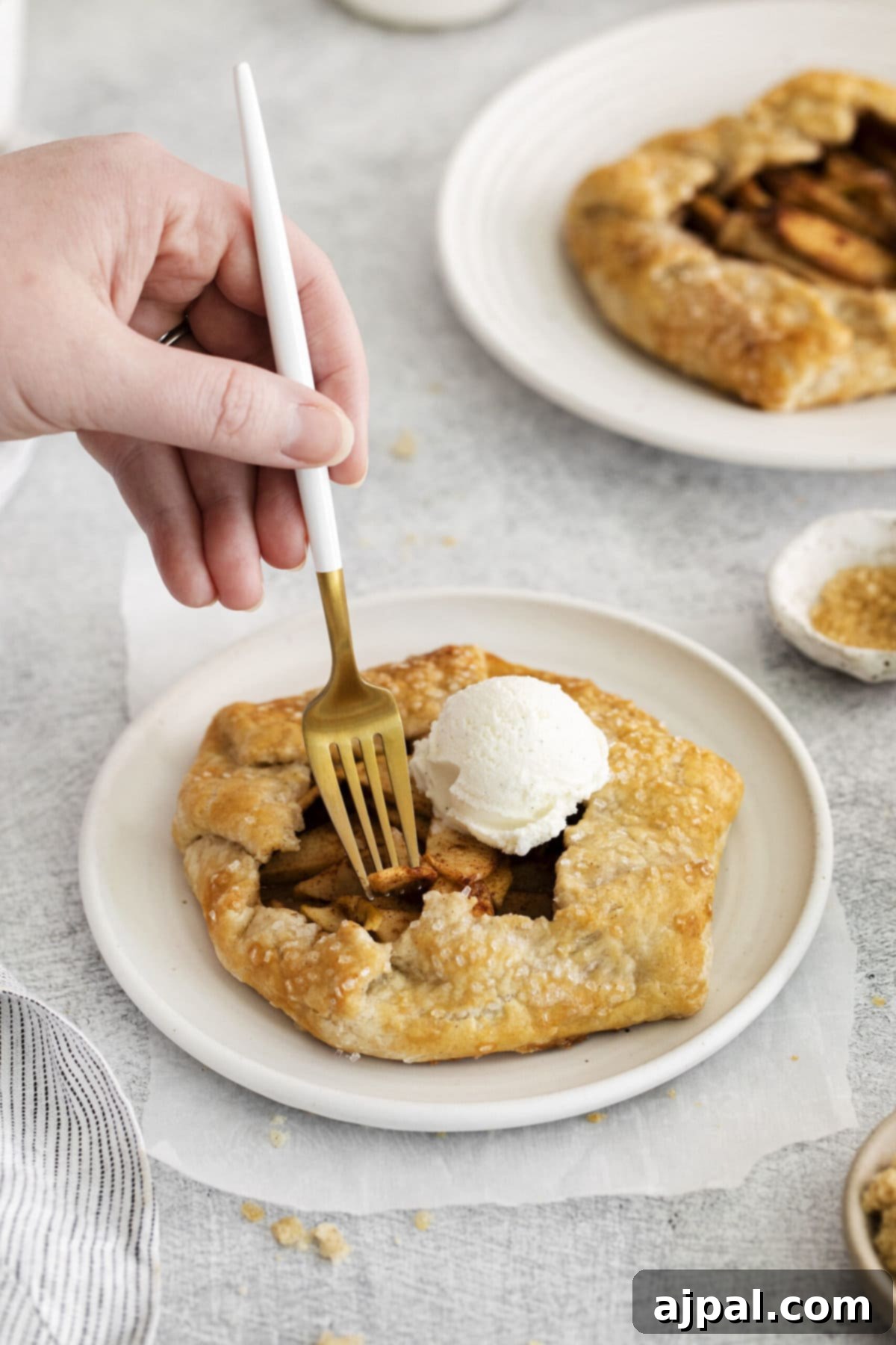 A hand with a fork cutting into a warm apple galette, revealing the soft apple filling and flaky crust, served with ice cream.
