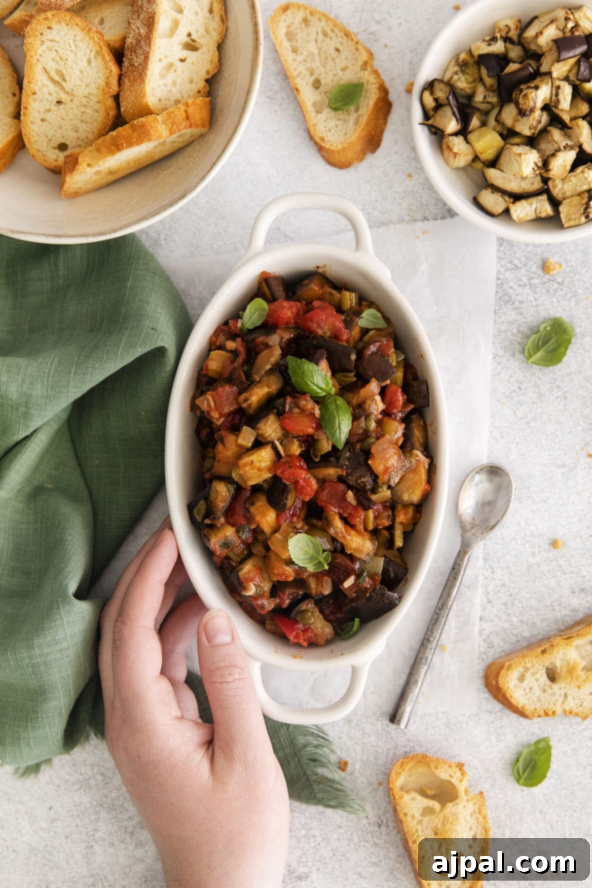 No-Fuss Eggplant Caponata 2 A beautifully baked eggplant caponata in a white serving dish, garnished with fresh basil leaves, resting on a rustic wooden table next to a silver spoon.