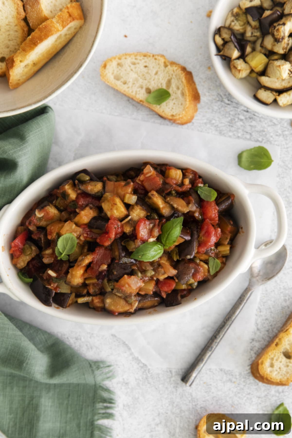 No-Fuss Eggplant Caponata 3 A cozy table setting featuring a white bowl brimming with homemade eggplant caponata, accompanied by slices of fresh, crusty bread.