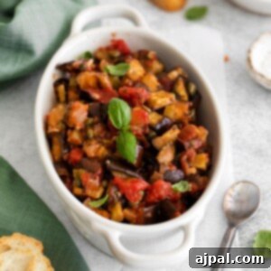 A picture of baked eggplant caponata in a white dish with basil leaves on top on a table next to a spoon.