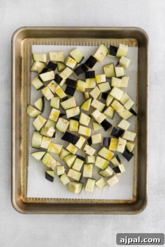 No-Fuss Eggplant Caponata 5 Evenly cubed raw eggplant pieces are neatly arranged on a baking sheet lined with parchment paper, glistening slightly with oil, ready for roasting.