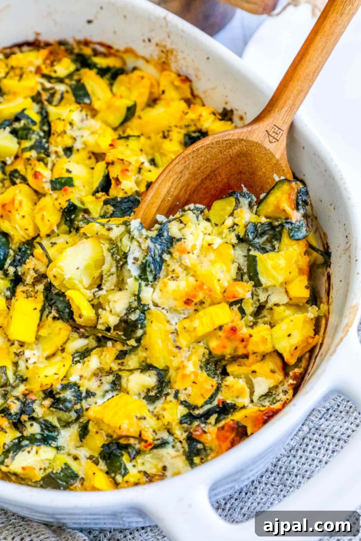 Green & Gold Feta Casserole 3 A spoon scooping a generous portion of warm, cheesy spinach, zucchini, and feta casserole from a white baking dish.