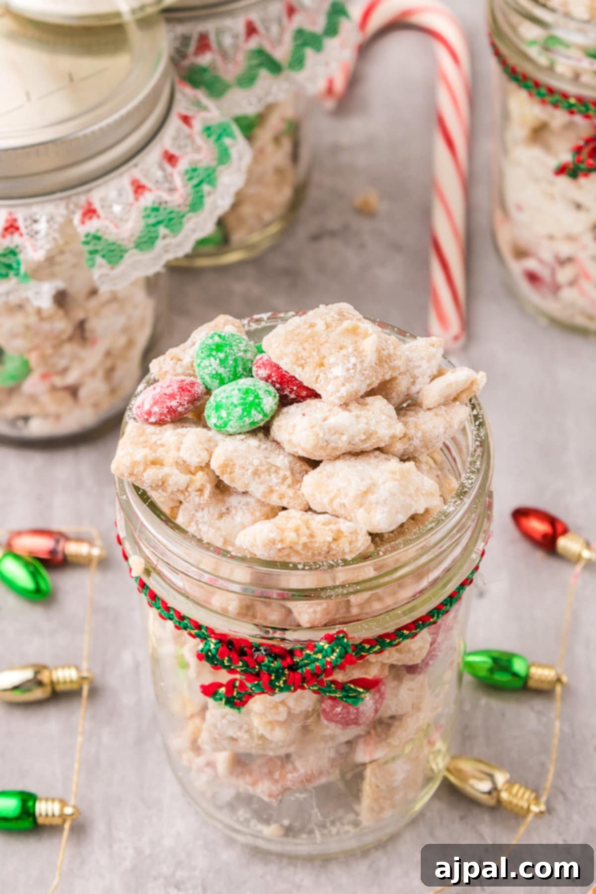 picture of muddy buddies chex mix with pretzels and red and green candies mixed in coated in powdered sugar in a jar with a red and green bow on it 
