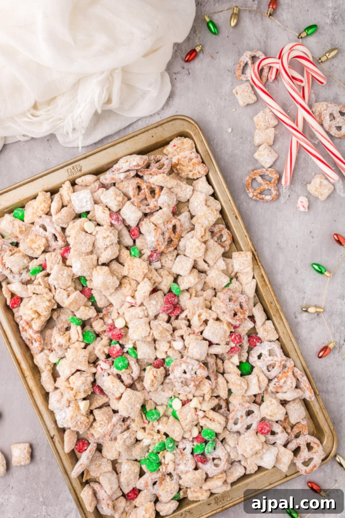 picture of muddy buddies chex mix with pretzels and red and green candies mixed in coated in powdered sugar on a baking sheet