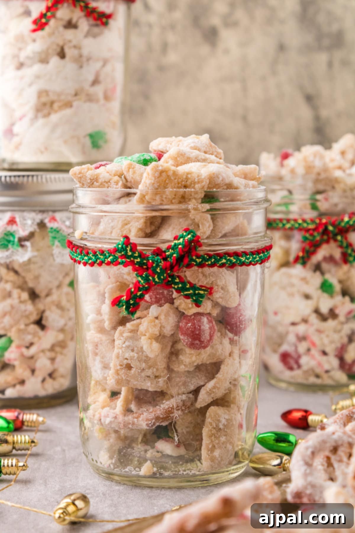 picture of muddy buddies chex mix with pretzels and red and green candies mixed in coated in powdered sugar in a jar with a red and green bow on it 
