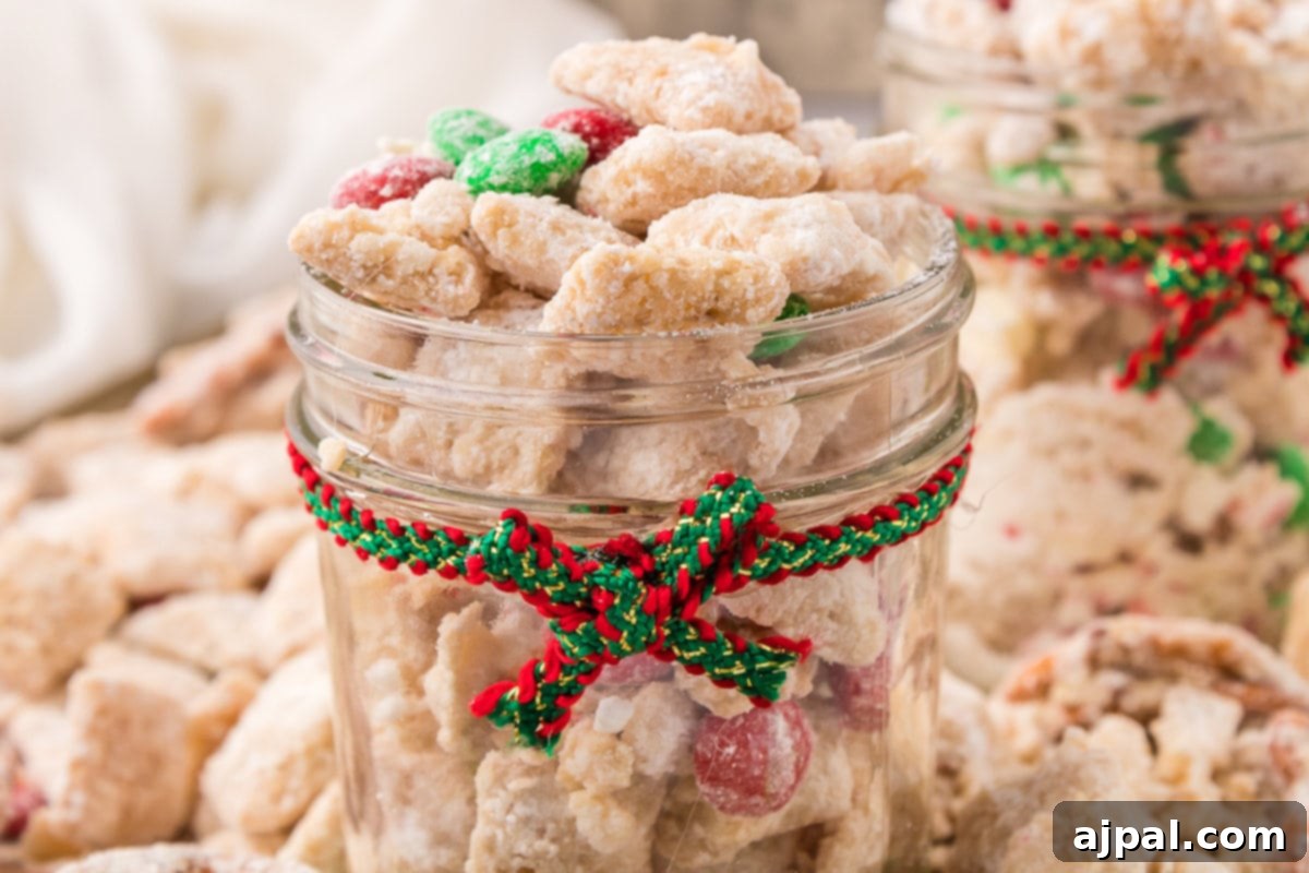 picture of muddy buddies chex mix with pretzels and red and green candies mixed in coated in powdered sugar in a jar with a red and green ribbon around it