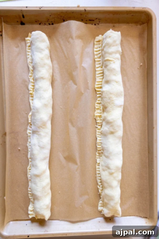 Classic English Sausage Rolls Recipe 8 Long, uncut sausage rolls resting on parchment paper, prepared and ready for slicing and baking.