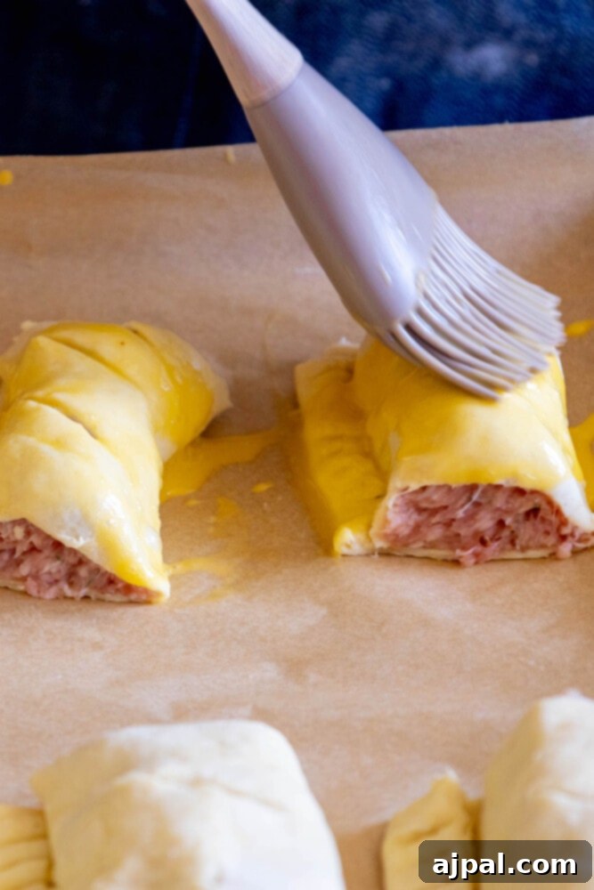 Classic English Sausage Rolls 9 Uncooked sausage roll being brushed with egg wash on parchment paper before baking.