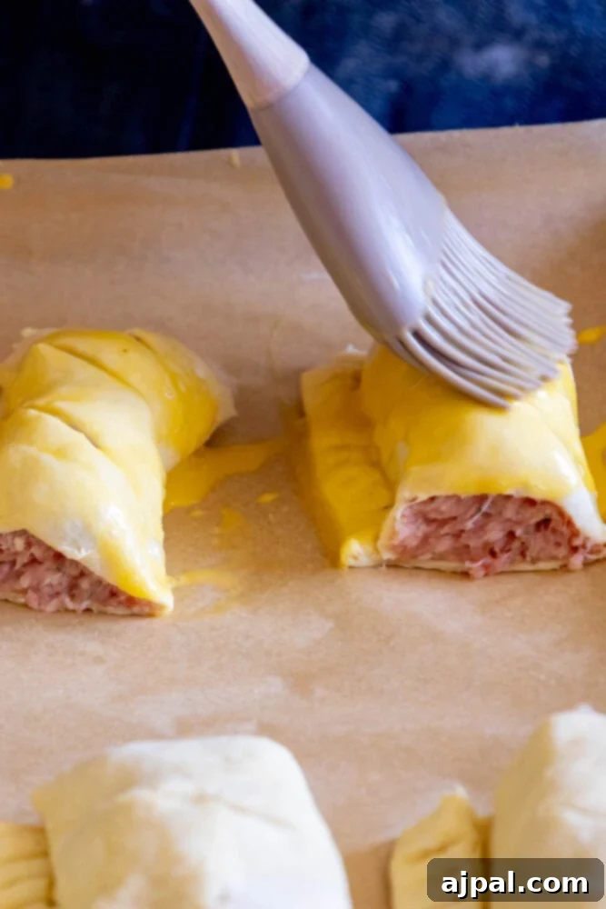 Classic English Sausage Rolls Recipe 9 An uncooked sausage roll being brushed with an egg wash on parchment paper, preparing it for a golden finish.