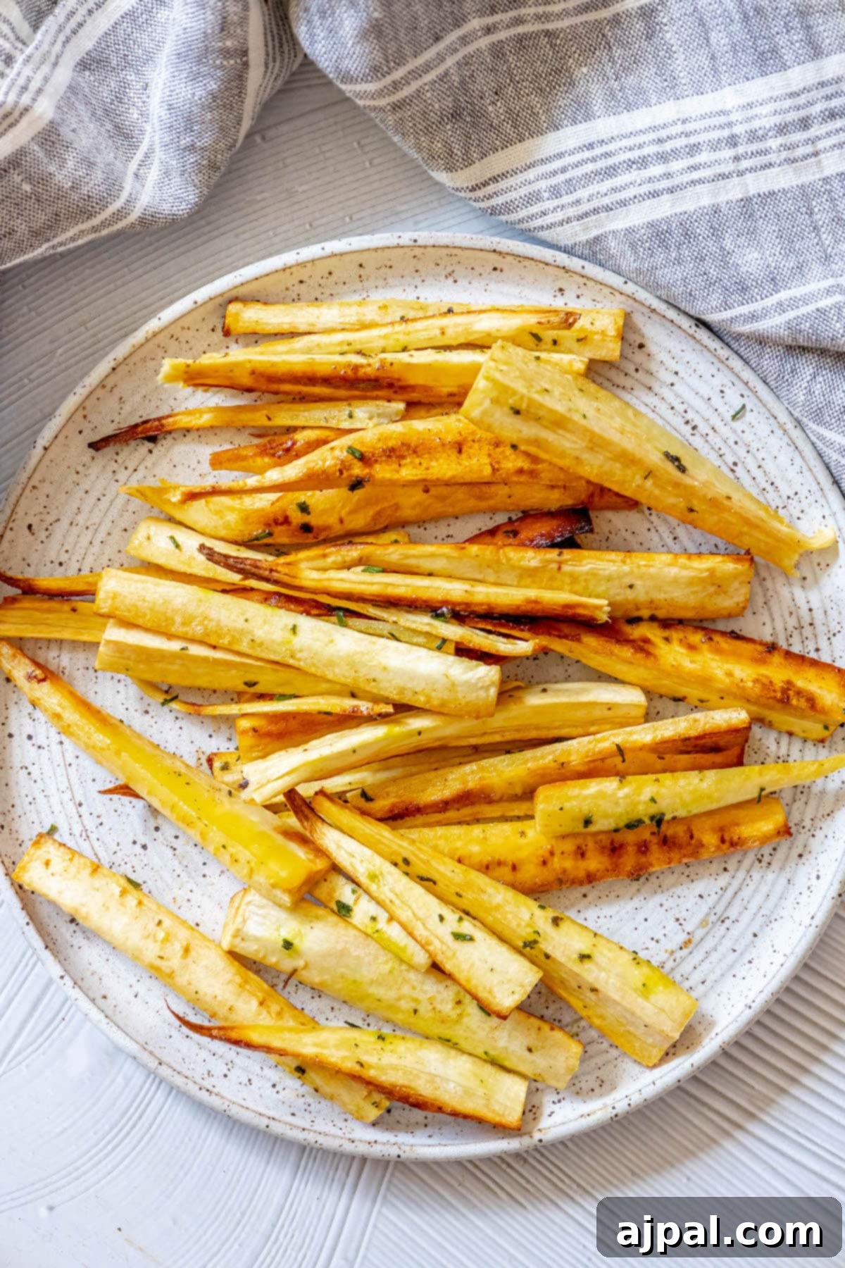 roasted parsnips seasoned with garlic butter and herbs on a white speckled plate next to a blue striped tea towel