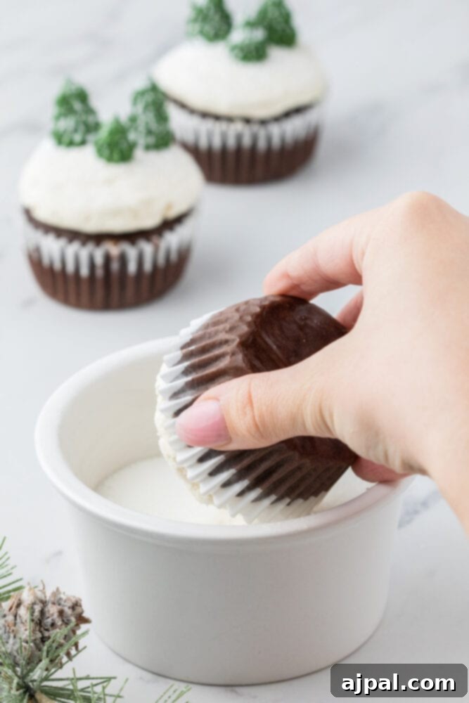 A hand gently dips a frosted cupcake into a bowl of fine powdered sugar, creating a beautiful 'snowy' topping for a winter-themed treat.