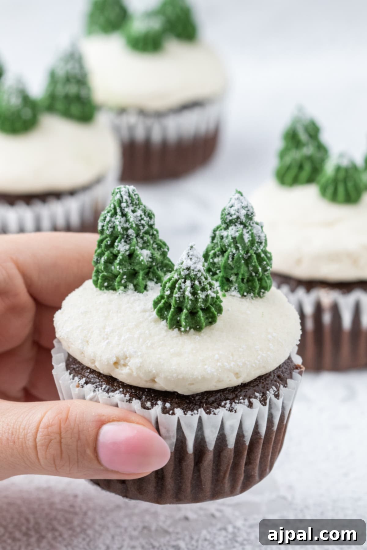 A hand gently holds a winter mint chip cupcake, showcasing its delicate buttercream icing and a piped Christmas tree atop a snowy powdered sugar base.