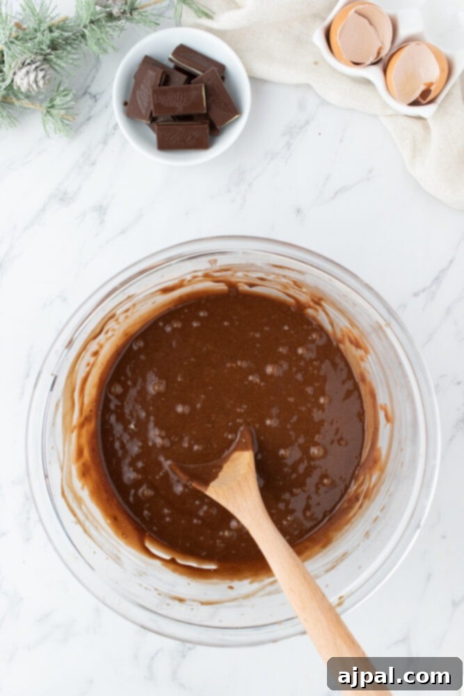 A rich chocolate mint cupcake batter being gently mixed in a glass bowl with a wooden spoon, resting on a marble-patterned table.