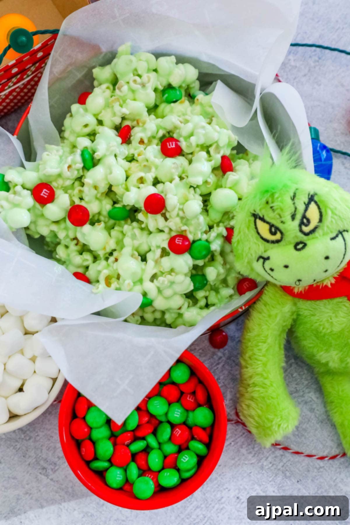 A festive bowl of green Grinch popcorn, adorned with colorful Christmas M&Ms, sits beside a charming Grinch doll, ready for a holiday movie night.