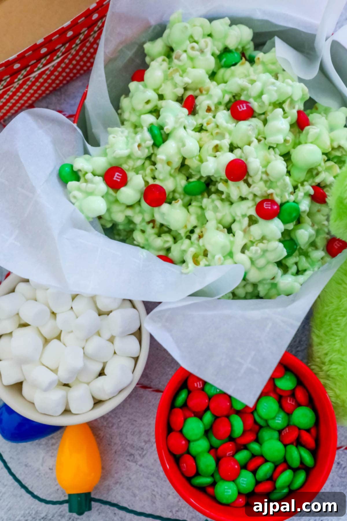 A bowl of green candy-coated popcorn with vibrant green and red Christmas candies, set on a table with twinkling Christmas lights and M&Ms, creating a cozy festive scene.