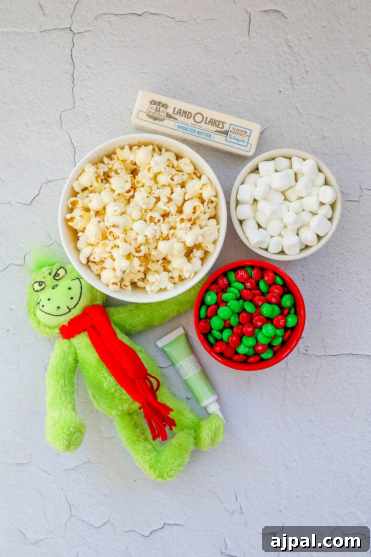 A collection of ingredients for Grinch popcorn: a Grinch doll, a bowl of popped popcorn, green food coloring, M&M candies, marshmallows, and butter, arranged on a rustic wooden table.