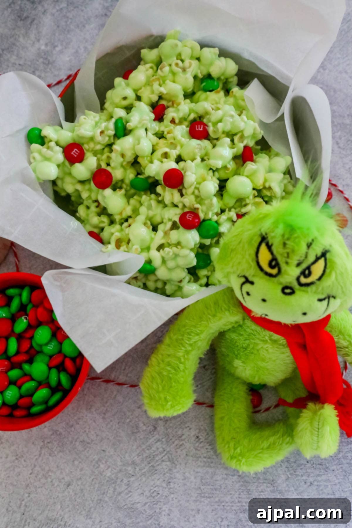 A bowl of green candied Christmas popcorn, with vibrant green and red M&Ms, sits beside a Grinch doll, surrounded by more M&Ms.