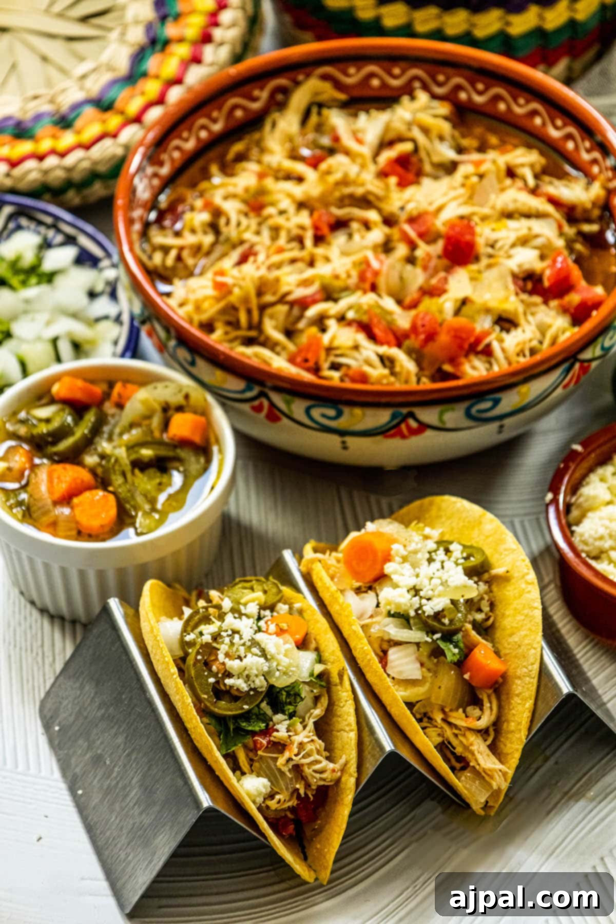 Shredded chicken with tomatoes and green chile in a beautiful talavera bowl, perfectly complemented by ready-to-eat chicken tacos in a taco stand. The table is adorned with vibrant side dishes including jalapeno and carrot salsa, crumbled white cheese, fresh limes, diced onions, and a stack of warm tortillas.