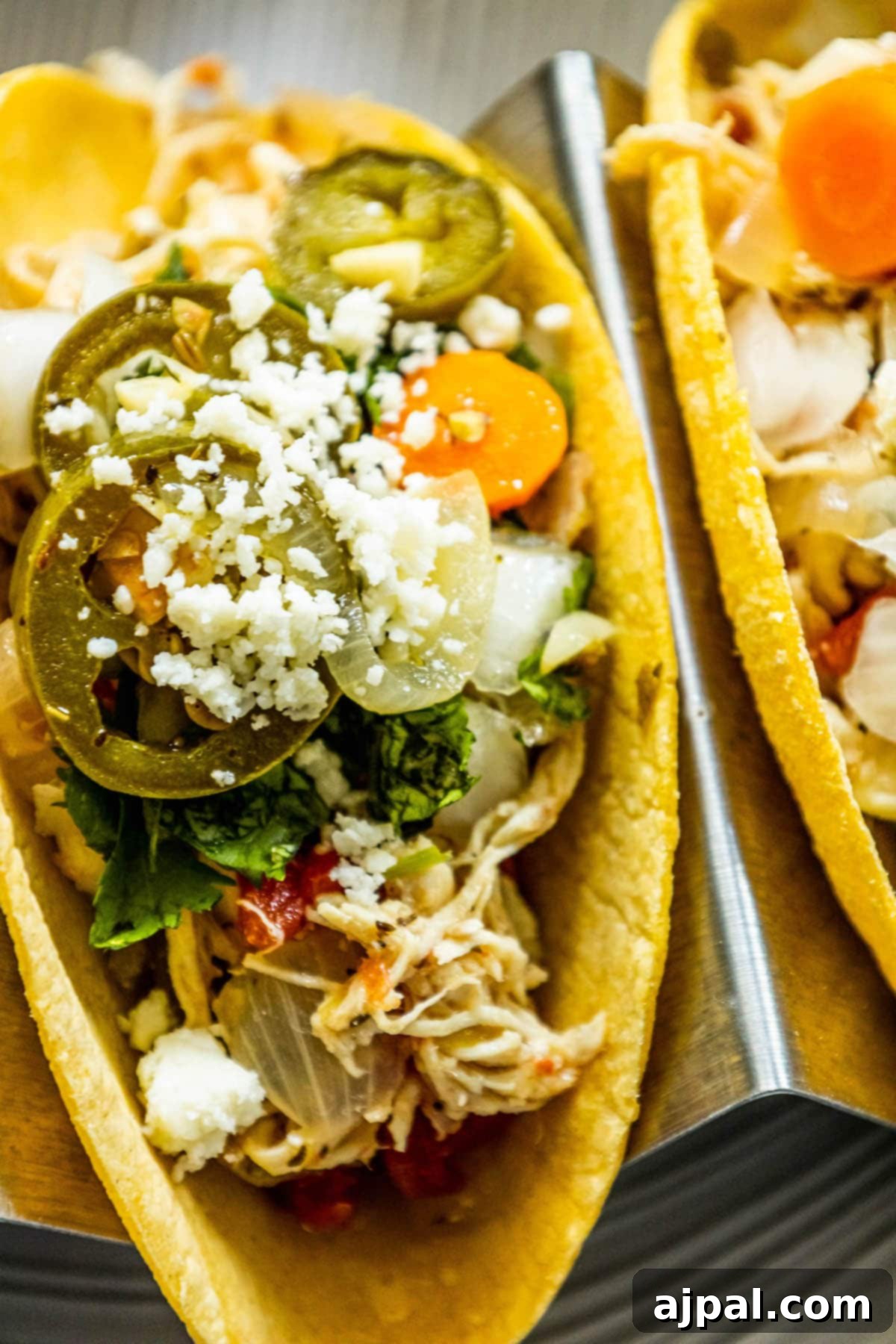 Two vibrant green chile shredded chicken tacos, meticulously arranged in a sleek metal taco stand. Each taco is generously filled with tender shredded chicken, tangy pickled jalapenos, rich crumbled white cheese, crisp diced onions, fresh chopped cilantro, and savory pickled carrots.
