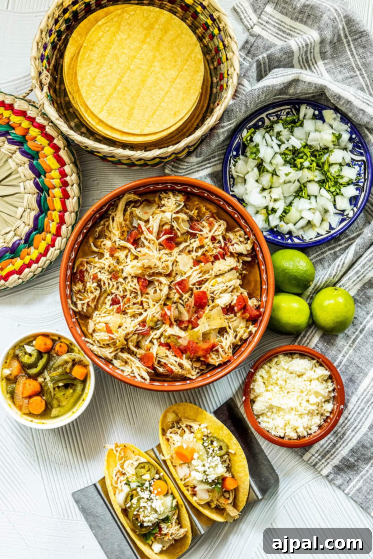 Shredded chicken with tomatoes and green chile in a talavera bowl next to chicken tacos in a taco stand and a bowl of jalapeno and carrot salsa, a bowl of crumbled white cheese, some limes, a bowl of diced onions, and a bowl of tortillas on a table