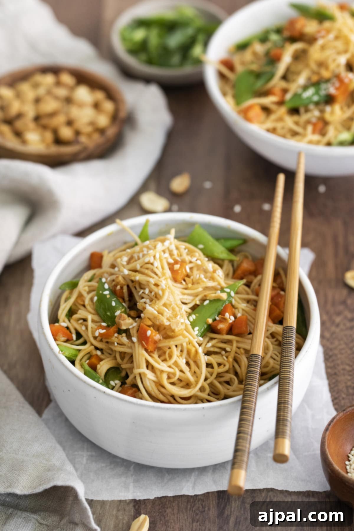 A bowl of spicy peanut noodles with carrots, snap peas, and chopped peanuts, served with chopsticks.
