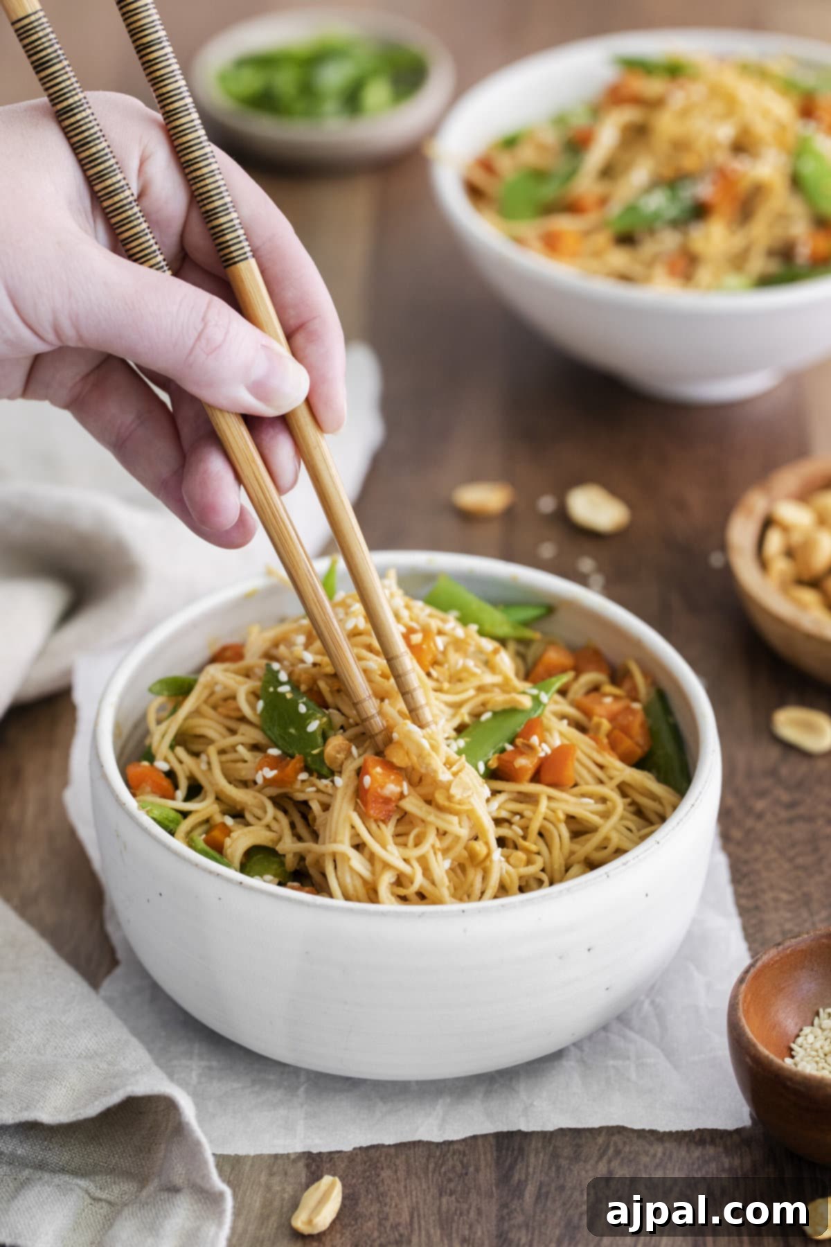 A bowl of vibrant peanut noodles with peas and carrots, being picked up with chopsticks.