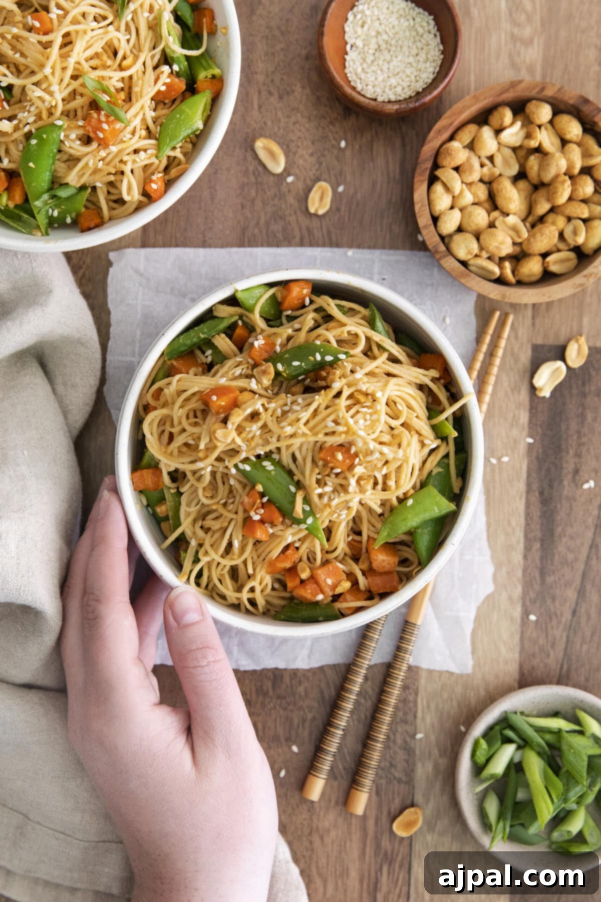 A hand holding chopsticks reaching for a bowl of thai noodles with carrots, peas, and a rich peanut sauce.