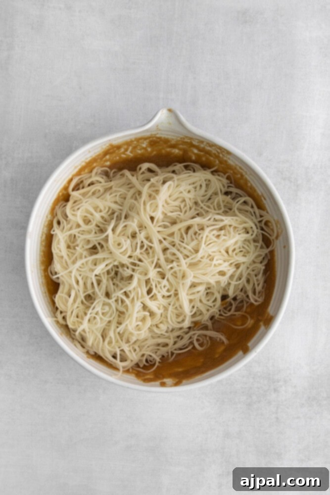 Cooked thin Asian noodles being coated in a spicy peanut sauce in a large mixing bowl.