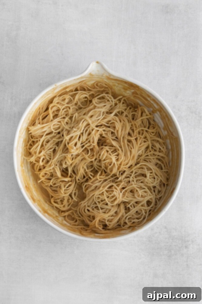 Close-up of thin Asian noodles fully coated in a rich peanut sauce in a mixing bowl.