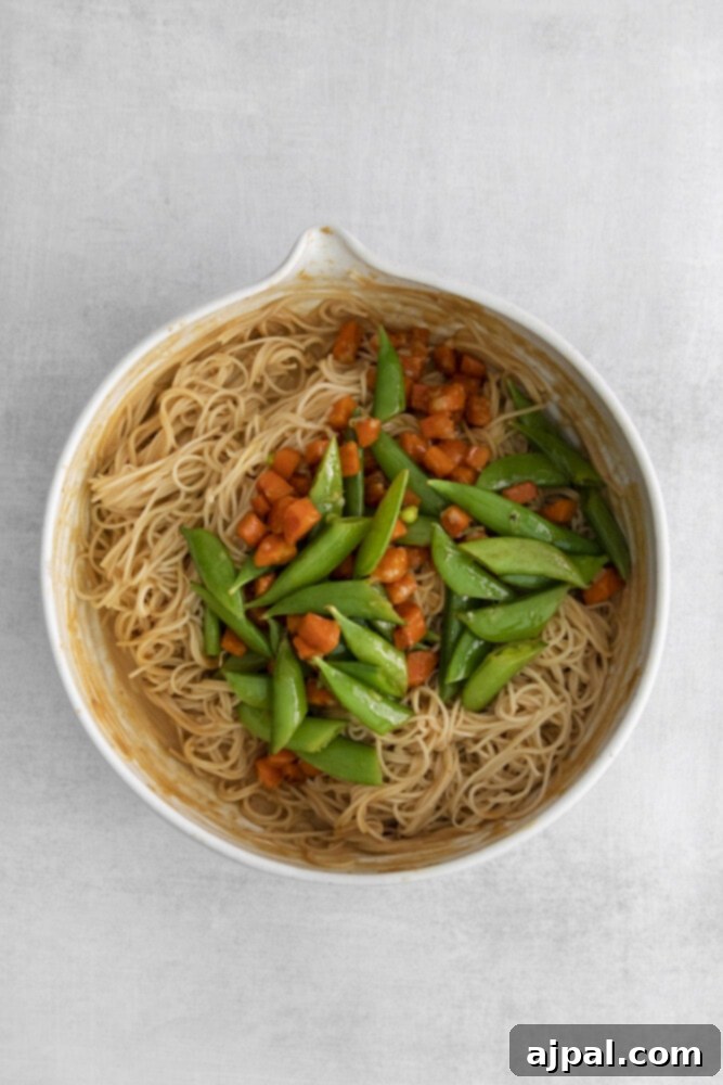 Snap peas and carrots being added to the peanut-coated noodles in a mixing bowl.