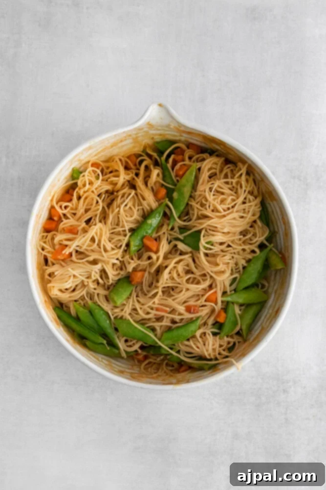 Kickin' Peanut Noodles 9 A close-up of noodles, tender snap peas, and crisp carrots being thoroughly mixed in a creamy, spicy peanut sauce, showcasing the vibrant colors and appealing texture of the dish.