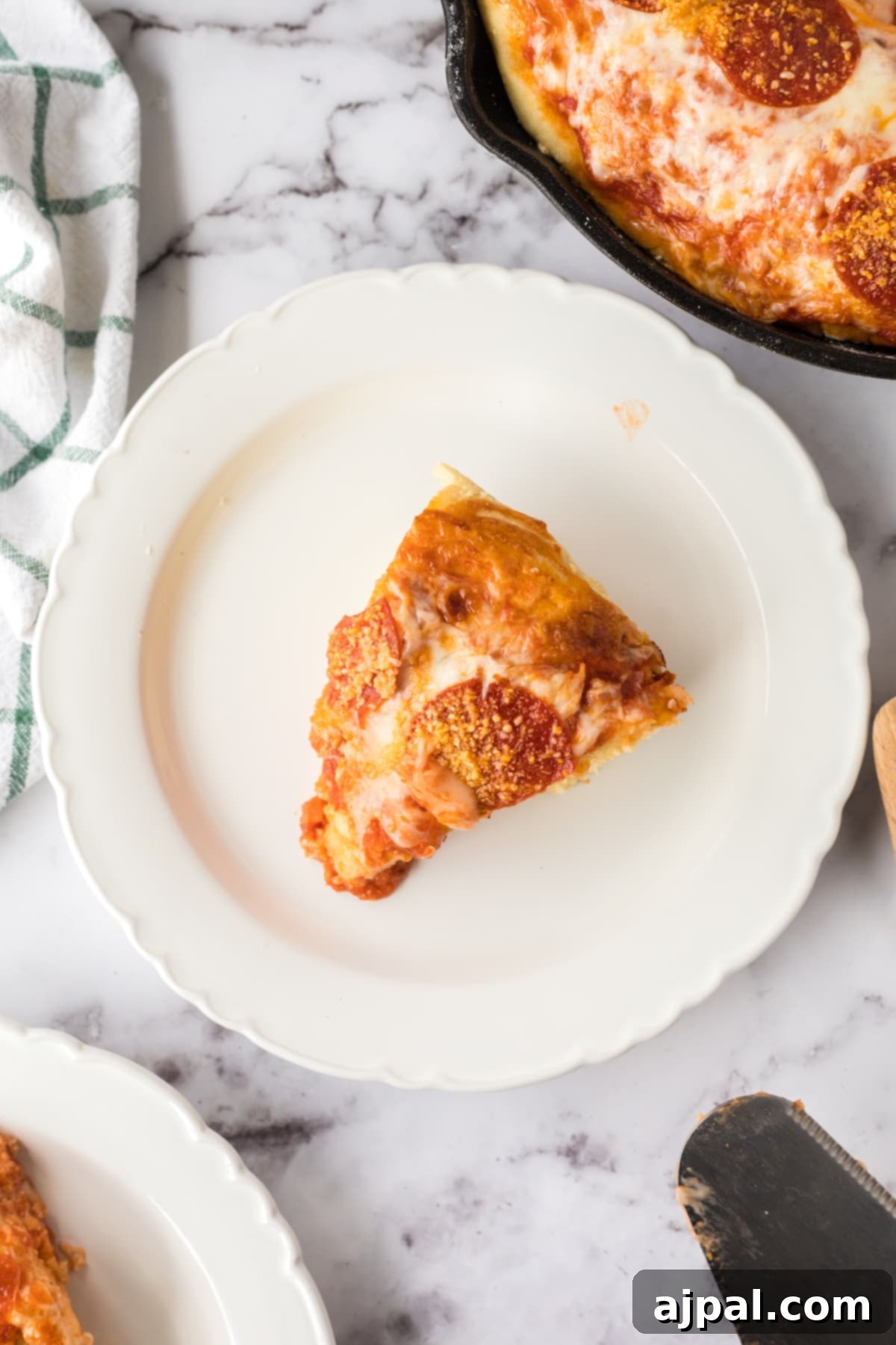 A delectable slice of deep dish pizza on a plate, positioned beside a cast iron skillet holding the rest of the freshly baked pizza.