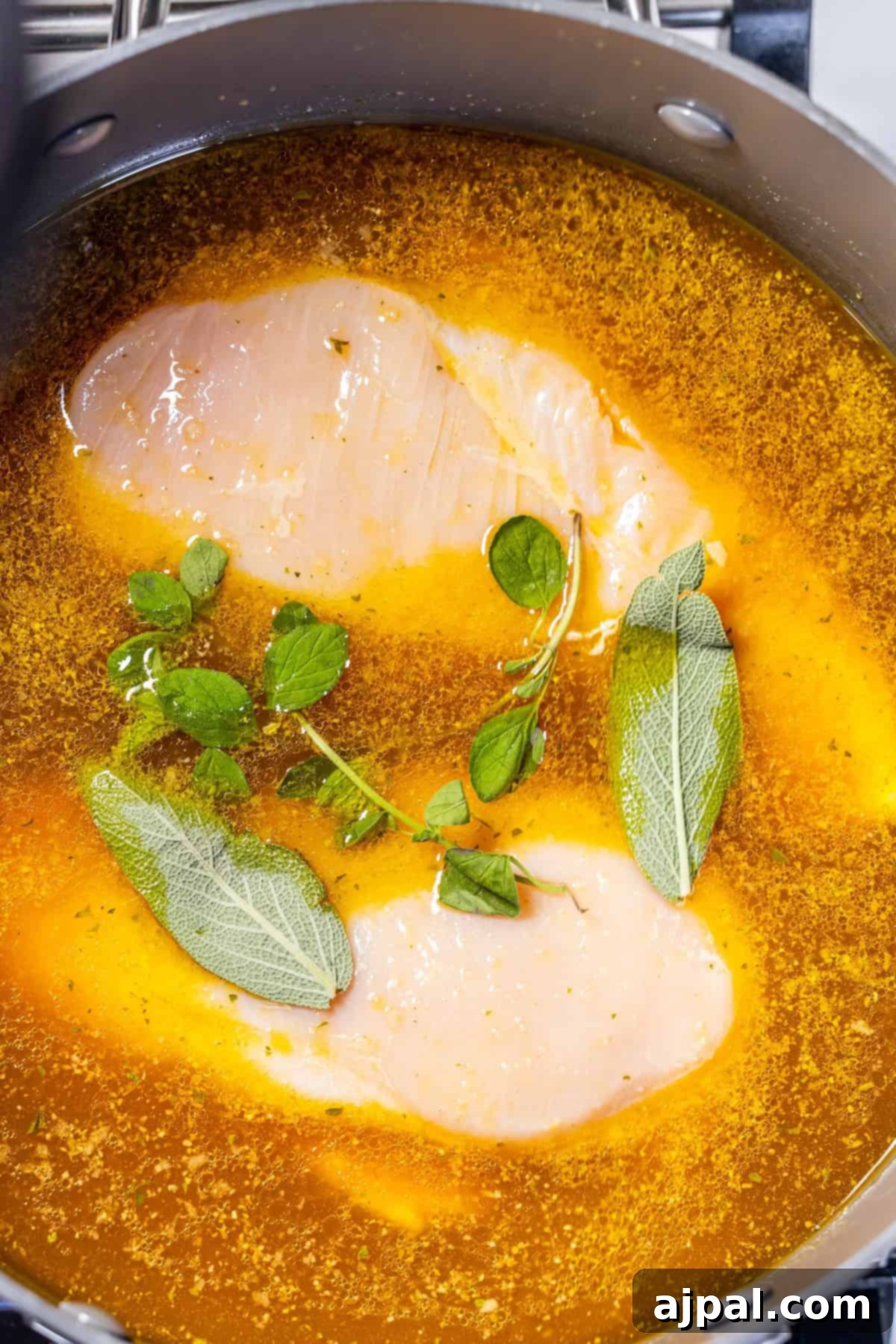 Zesty Green Chicken Posole 6 Two raw chicken breasts simmering in a pot with an orange-hued broth, garnished with fresh sage and oregano. This illustrates the initial boiling step for tender chicken, ideal for a green chicken posole recipe.