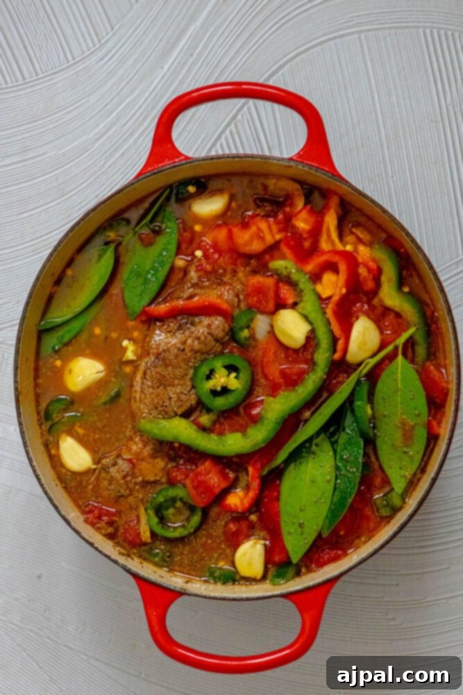 Spicy Mexican Pot Roast 10 A pot roast nestled in a Dutch oven, surrounded by sliced bell peppers, jalapeños, garlic cloves, and bay leaves, all simmering in beef stock.