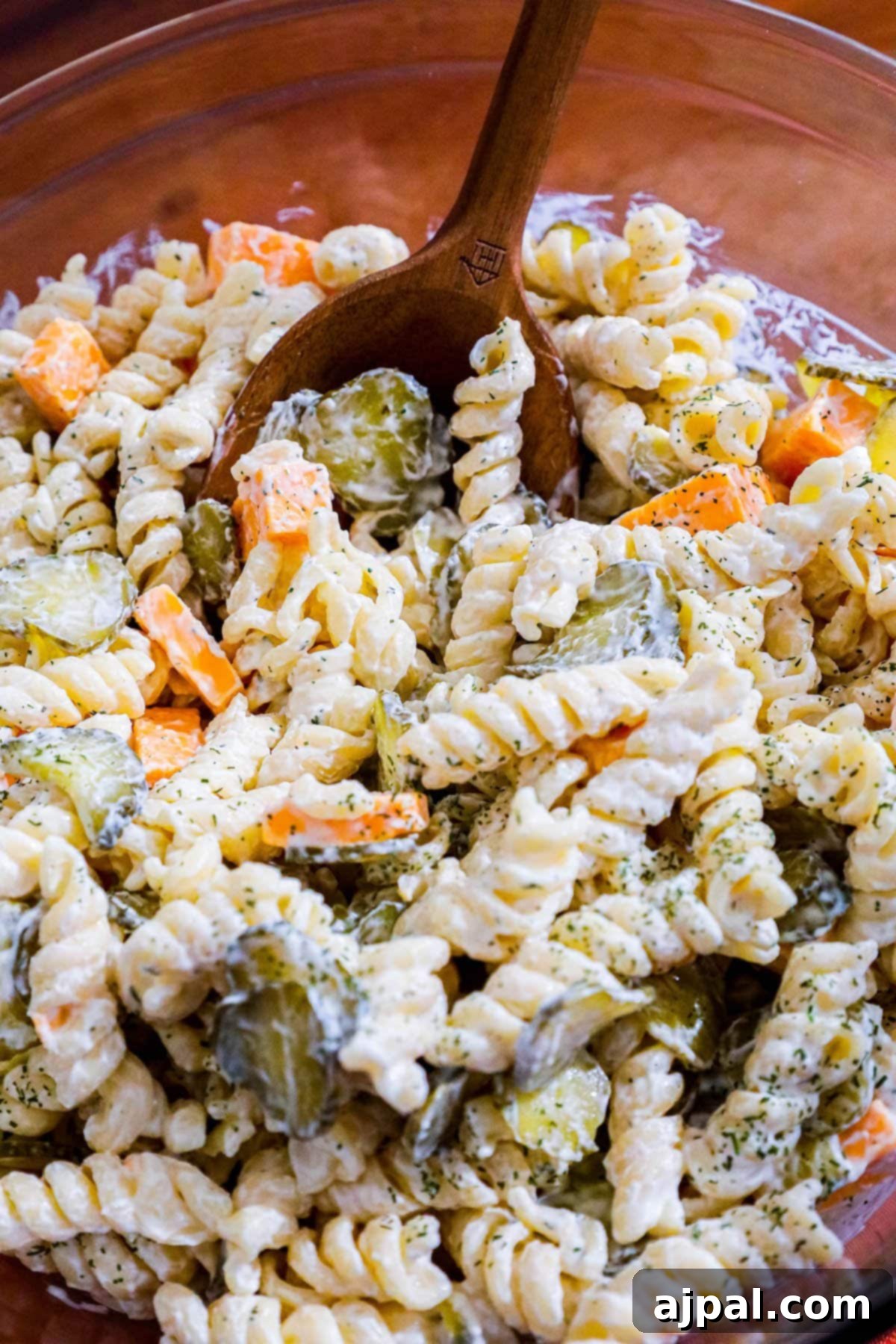 Tangy Dill Pasta Delight 6 A wooden spoon scooping out a portion of dill pickle pasta salad from a glass bowl, highlighting the creamy texture and pickle pieces.