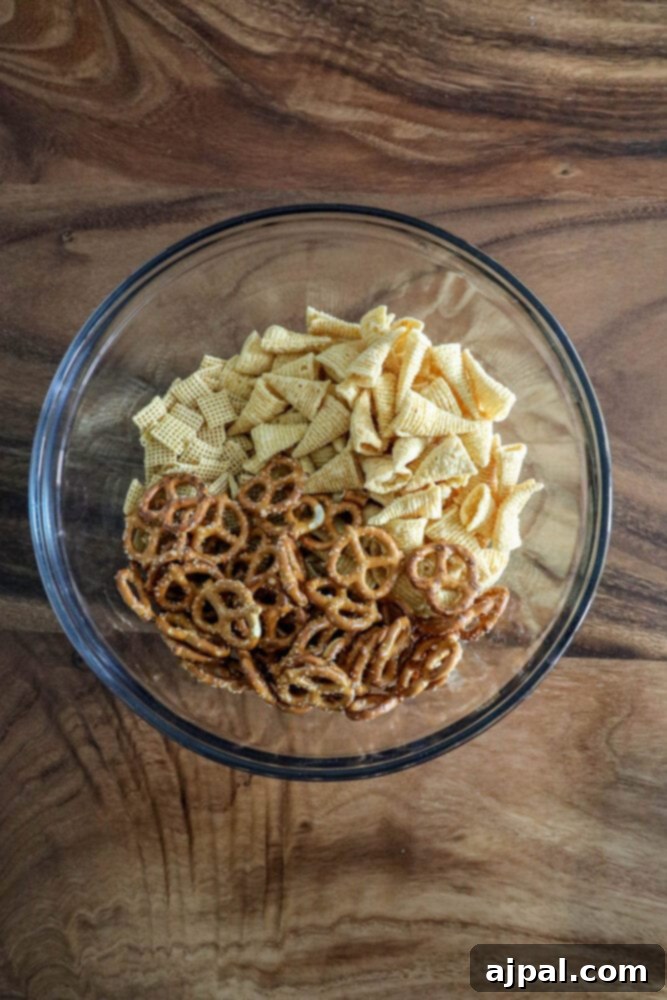 A clear glass bowl filled with the base ingredients for Chex Mix: corn chips, pretzels, and corn cereal, ready for seasoning.