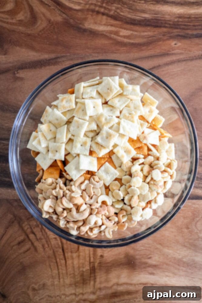 A clear glass bowl showcasing a mix of corn chips, corn cereal, pretzels, cheese crackers, and various nuts, forming the base of a savory snack.