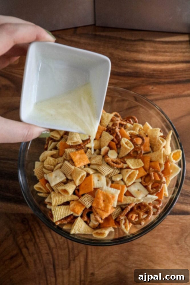 A hand gently pouring melted butter over a delightful mix of pretzels, corn chips, crackers, and corn cereal in a large mixing bowl, ensuring even coating.