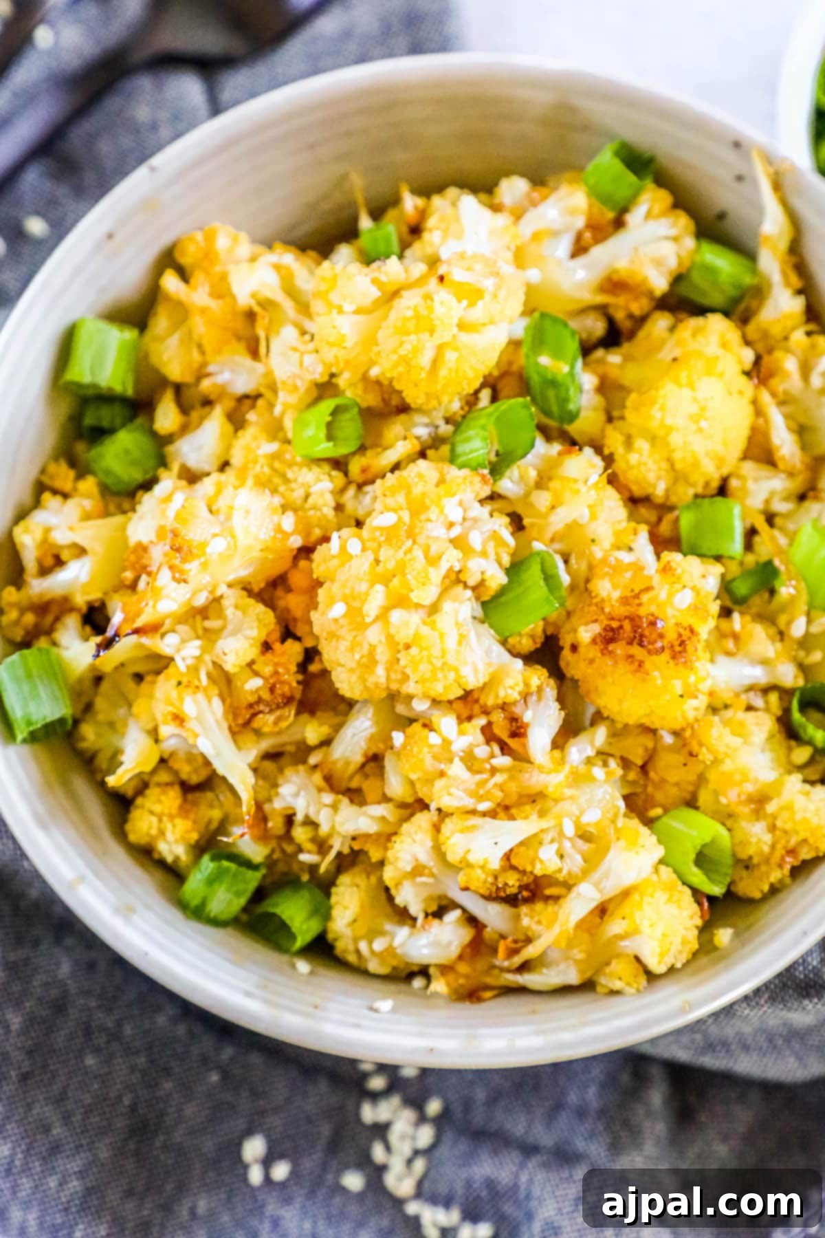 A beautifully baked cauliflower dish, garnished with toasted sesame seeds and vibrant sliced green onions, served in a pristine white bowl on a rustic wooden table. The cauliflower florets are perfectly browned and coated in a glistening, savory-sweet glaze.