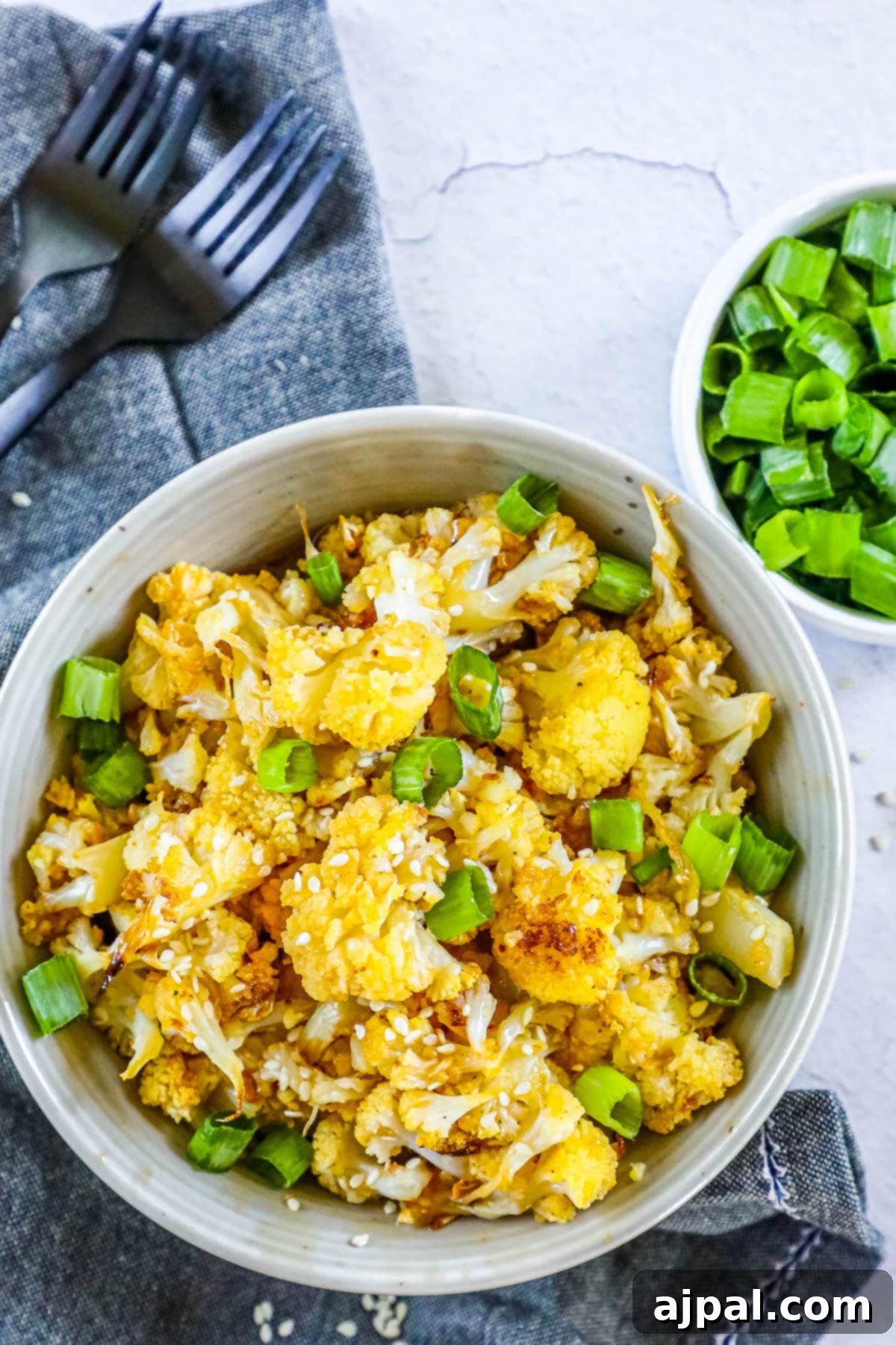 A delicious bowl of sweet and spicy baked cauliflower, topped with fresh green onions and sesame seeds, presented on a wooden table.