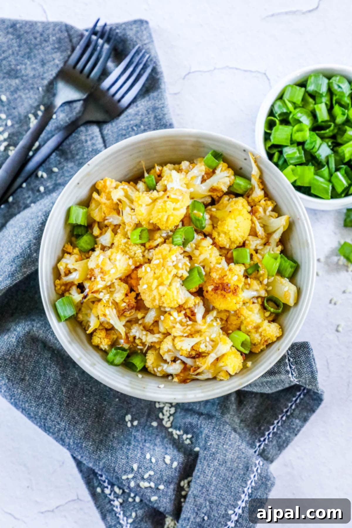 A close-up shot of sweet and spicy baked cauliflower, topped with fresh green onions and sesame seeds, presented in a stylish white bowl with forks nearby on a wooden table, ready to be enjoyed.