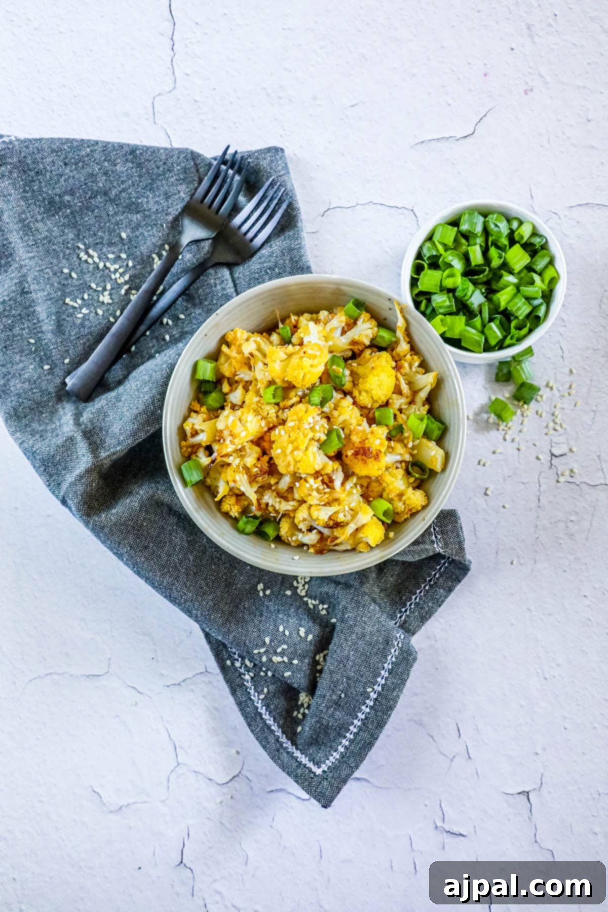 A serving of sweet and spicy baked cauliflower, generously topped with fresh green peas and sesame seeds, presented in a white bowl on a wooden surface.