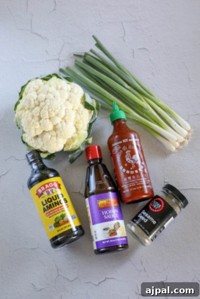 Fresh cauliflower florets, sriracha, hoisin sauce, liquid aminos, sesame seeds, and green onions neatly arranged on a table, showcasing the key ingredients for the recipe.